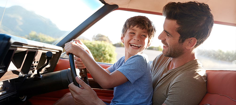image of a dad with his son sitting on his lap in the driver’s seat of a car.